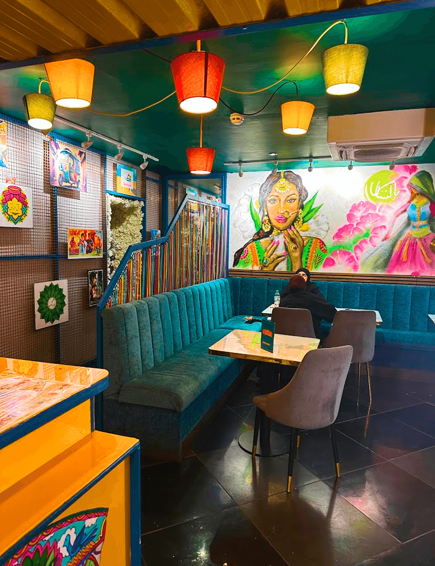 Restaurant interior with vibrant Pakistani atmosphere, teal booth seating, pendant lights, and traditional mural